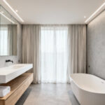 A serene, high-key minimalistic bathroom featuring light grey seamless walls, a floating oak vanity, a frameless mirror, and a standalone soaking tub, emphasizing simplicity to reduce cognitive load.