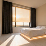 A bright, biologically optimized bedroom layout showing warm morning sunlight streaming across the bed, highlighting the importance of bed positioning near the light source and the presence of heavy blackout curtains for nighttime melatonin release.