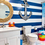 A full view photograph of a coastal bathroom overwhelmed by thematic kitsch, including rope-wrapped mirrors, bright blue stripes, and a large anchor decal above the toilet, demonstrating mass-produced theme decor.