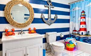 A full view photograph of a coastal bathroom overwhelmed by thematic kitsch, including rope-wrapped mirrors, bright blue stripes, and a large anchor decal above the toilet, demonstrating mass-produced theme decor.