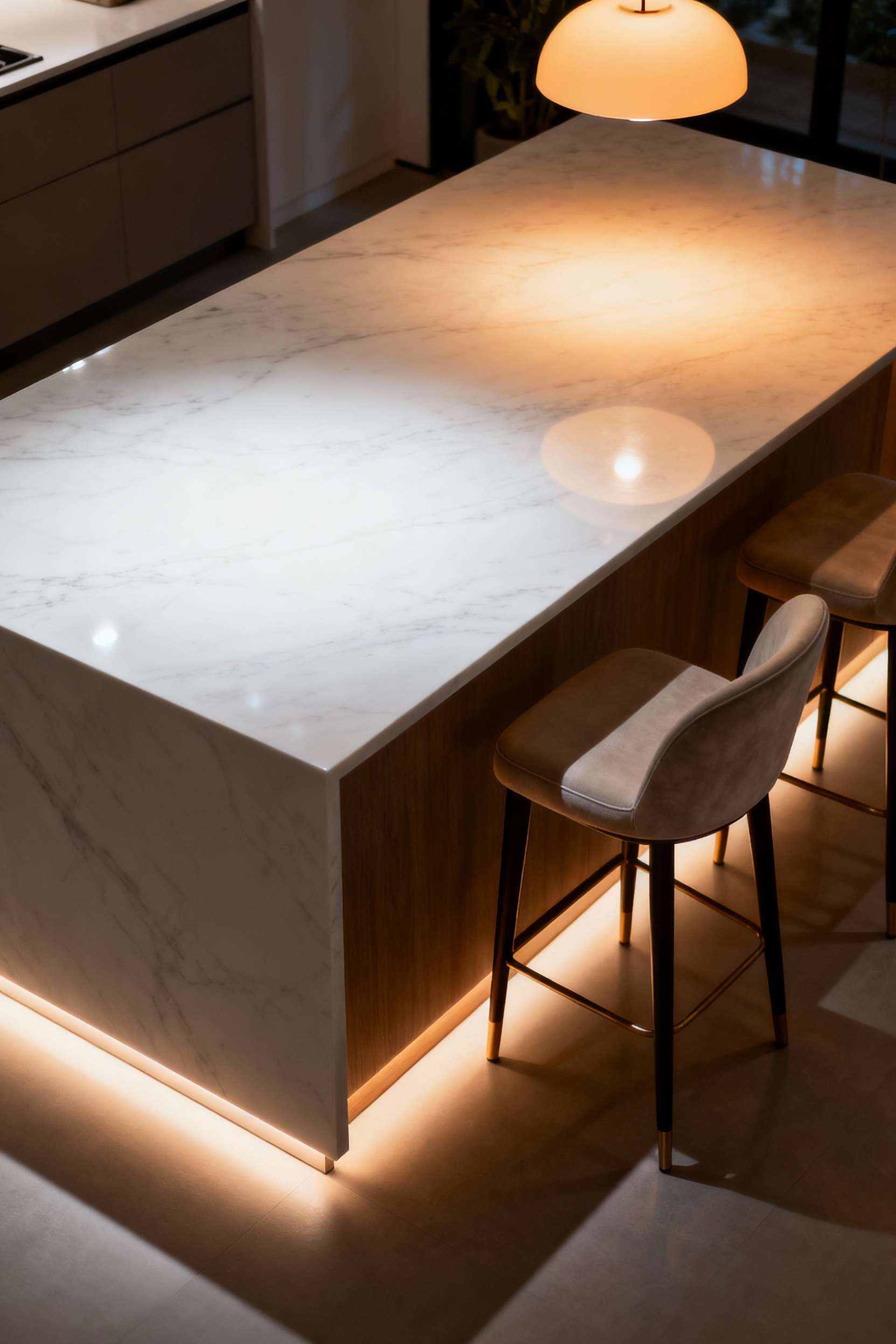 A beautifully lit modern kitchen island showcasing different lighting layers: bright task lighting over a marble prep area, warm ambient pendant light over a dining section, and soft toe-kick lighting at the base, demonstrating primary, secondary, and tertiary functions.