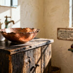 A visual distinction between authentic time-worn elegance (reclaimed wood vanity and hammered copper sink) and cheap, imitation kitsch accessories in a sunlit rustic bathroom.