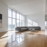 A minimalist modern living room with hardwood floors, a gray sectional sofa, and no area rug, illustrating a space that feels empty and echoing.